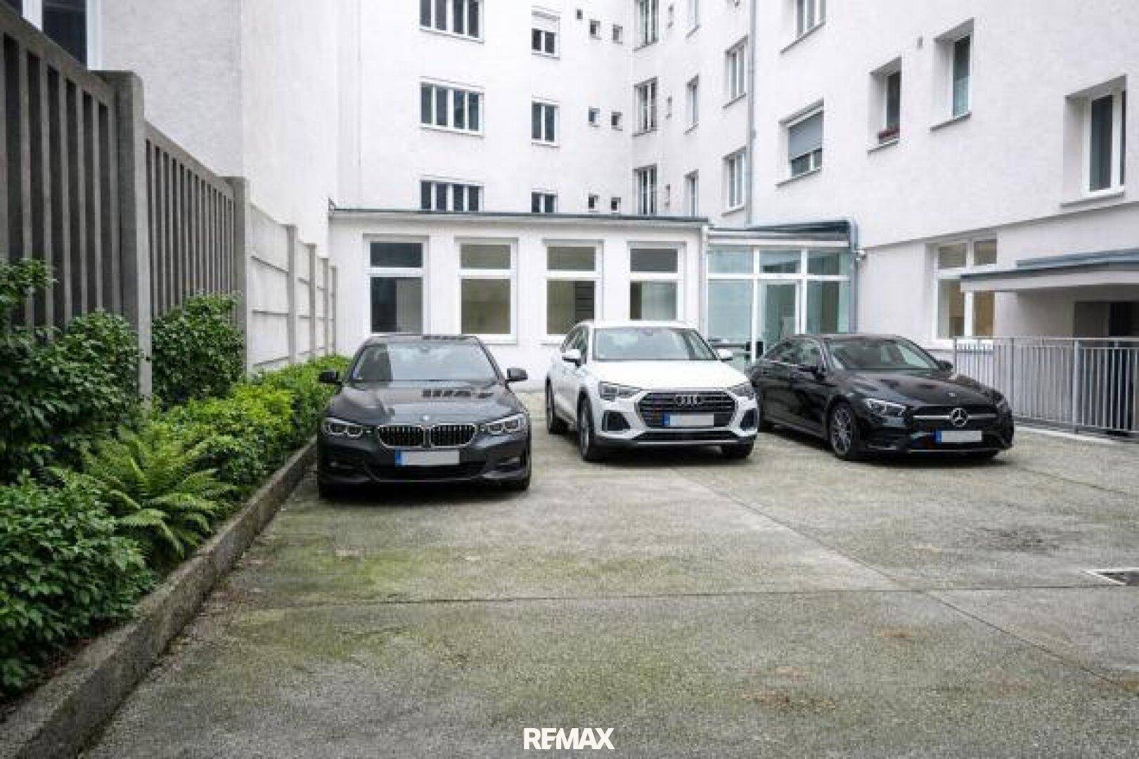Außenparkplatz mit drei geparkten Autos, umgeben von Gebäuden und etwas Grün.