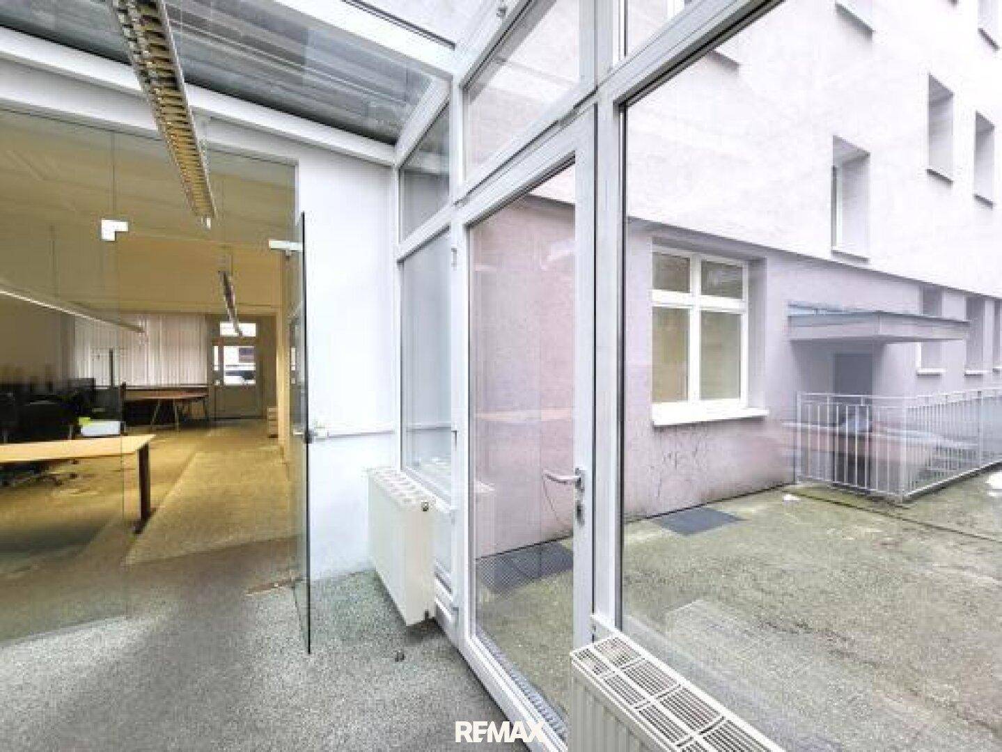 Blick aus einem verglasten Bereich auf einen kleinen Innenhof mit Gebäudefassaden.