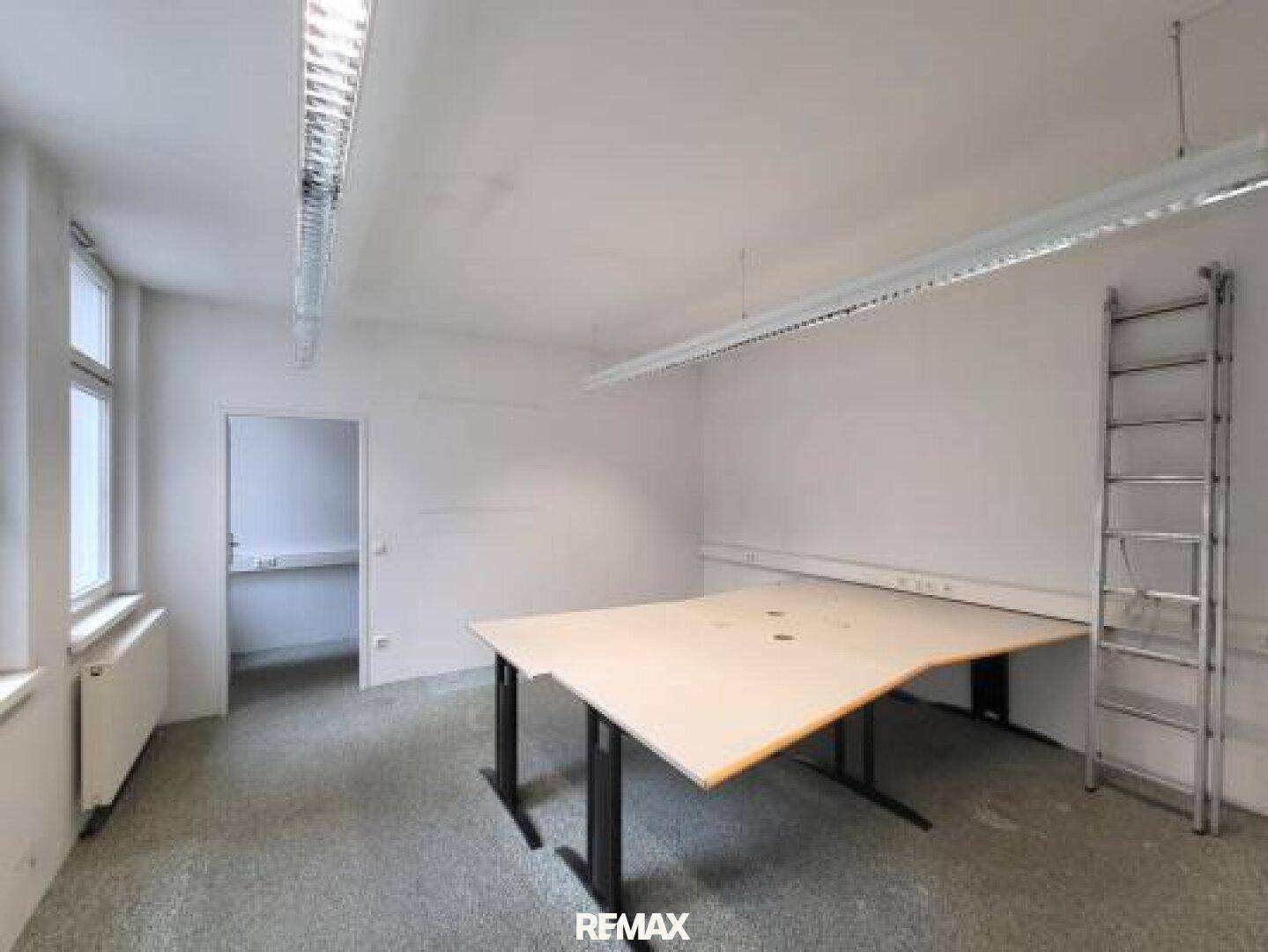 Leerer Büroraum mit großem Tisch, Fenstern und einer Leiter an der Wand.