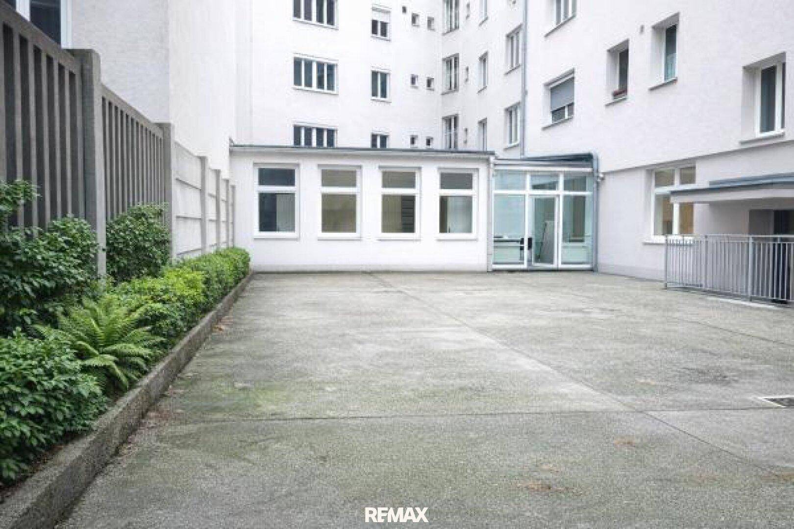 Leerer Außenparkplatz mit Betonfläche, umgeben von Gebäuden und einer Hecke.