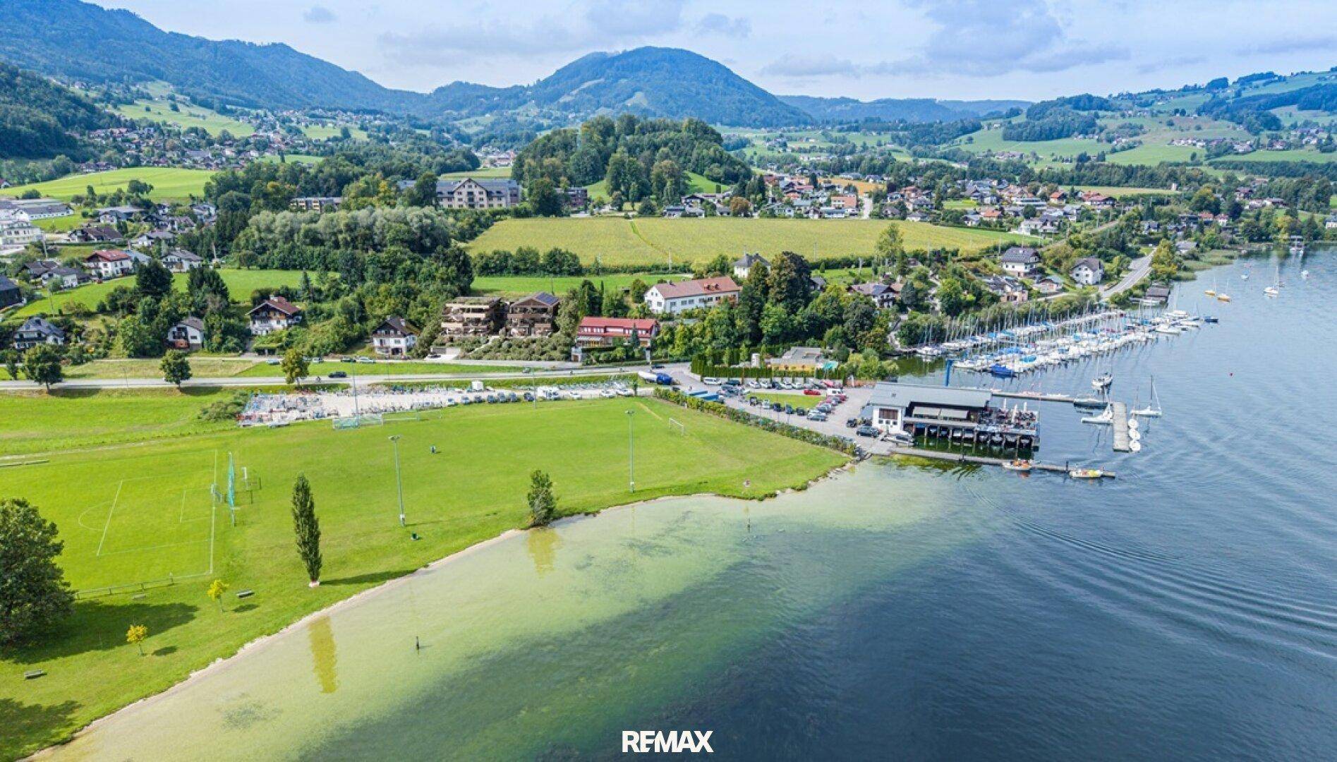 Luftaufnahme einer malerischen Seenlandschaft mit angrenzendem Dorf, Bergen und einem Yachthafen.