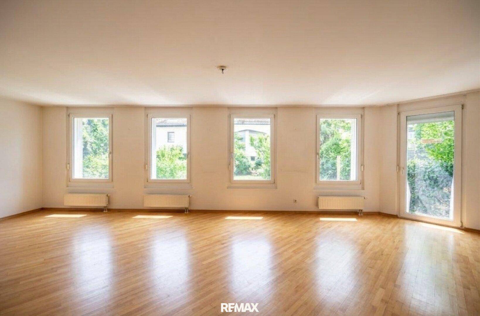 Helles, leeres Wohnzimmer mit Parkettboden und mehreren Fenstern, die viel Tageslicht hereinlassen.