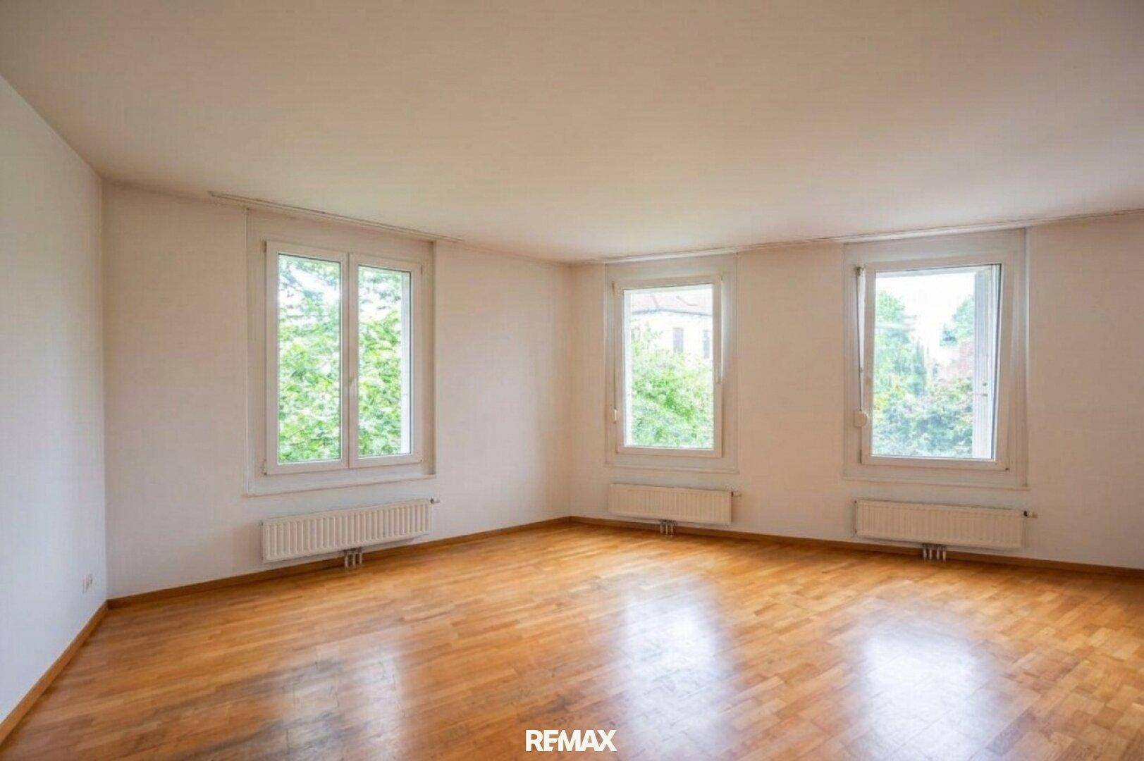 Geräumiges, leeres Wohnzimmer mit Holzboden und großen Fenstern, die eine grüne Aussicht bieten.