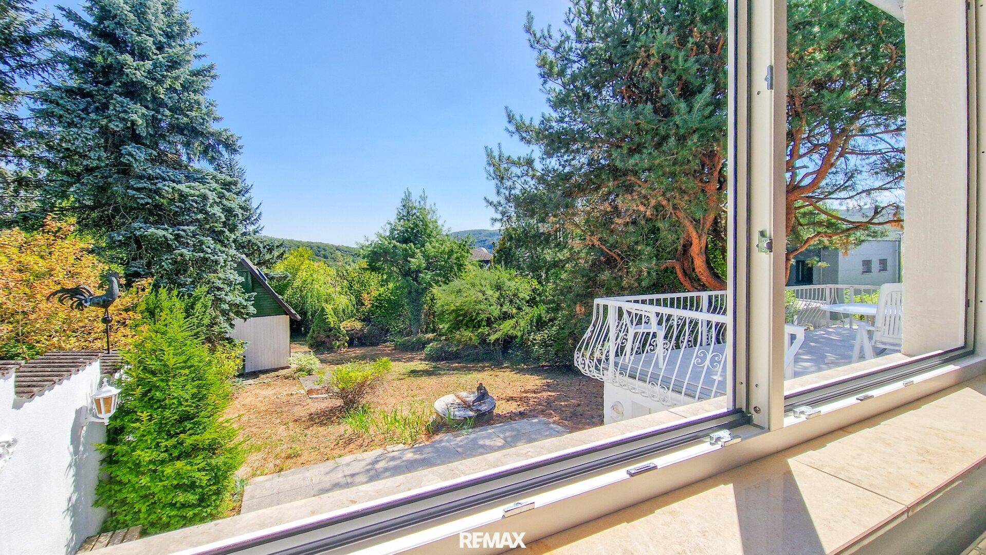 Blick aus dem Fenster auf einen weitläufigen Garten mit Bäumen und eine malerische Hügellandschaft im Hintergrund.