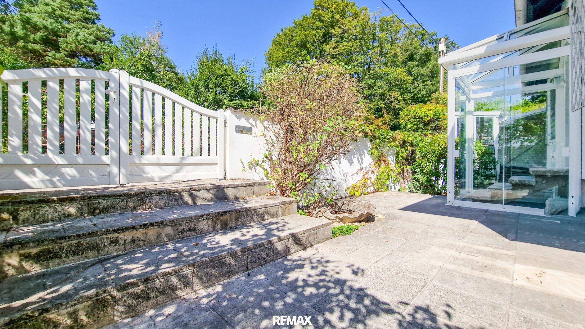 Steintreppe führt zu einer gepflasterten Terrasse mit moderner Glasstruktur und üppigem Grün.