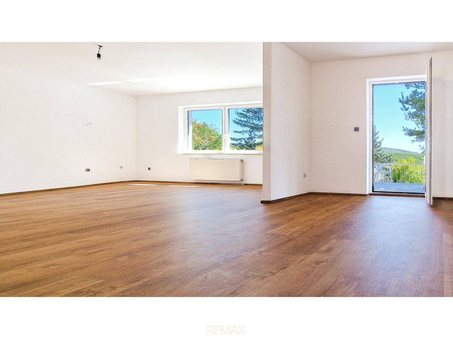 Großzügiger Wohnraum mit Holzboden und großen Fenstern, die viel Tageslicht hereinlassen und eine grüne Aussicht bieten.