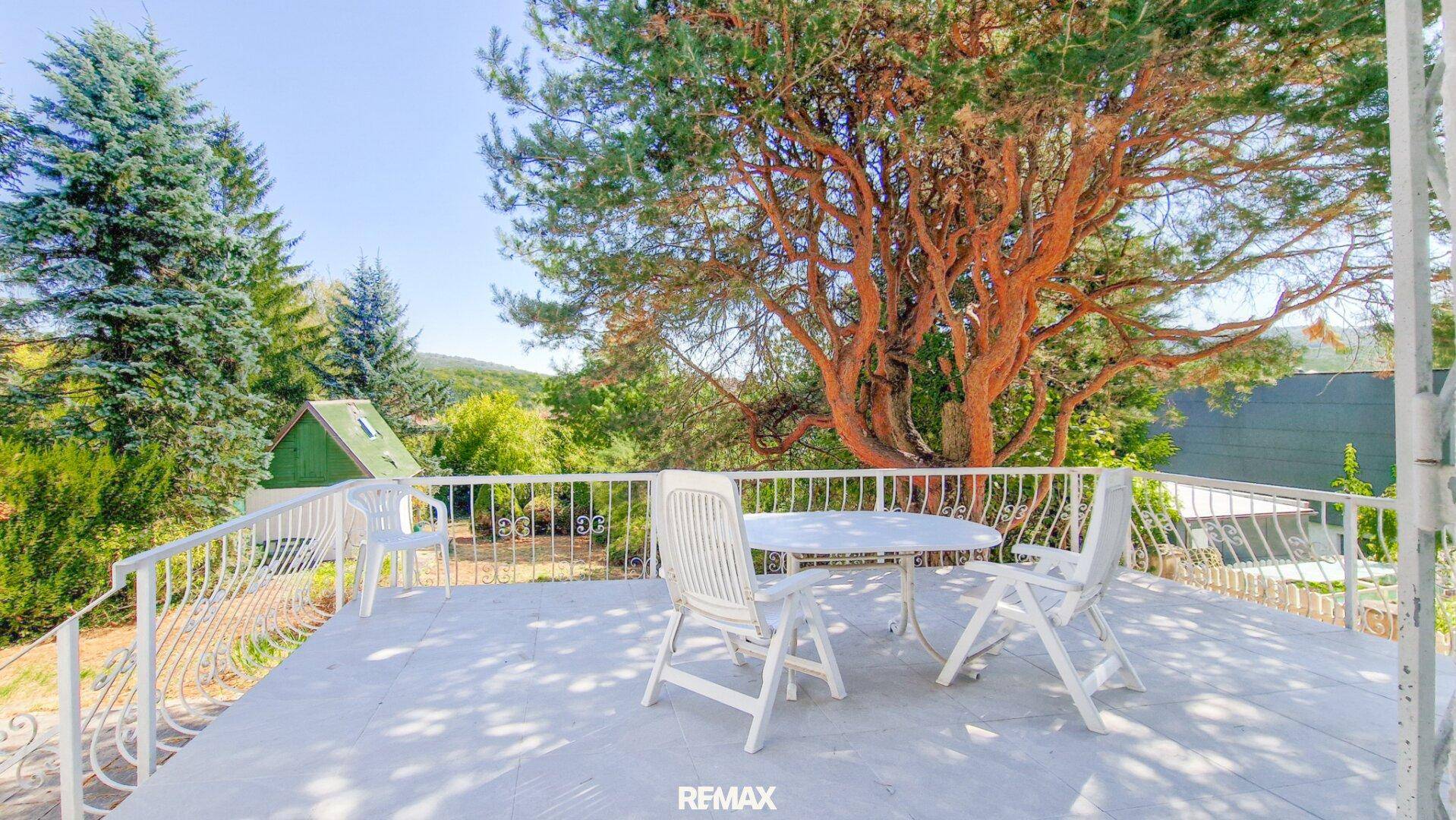 Sonnige Terrasse mit weißen Gartenmöbeln und schmiedeeisernem Geländer, umgeben von hohen Bäumen.