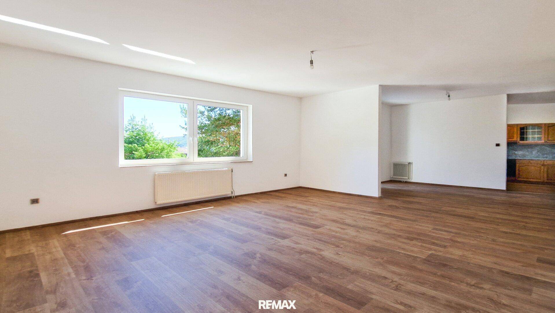 Geräumiges Wohnzimmer mit Holzboden und Fenster, das in einen angrenzenden Küchenbereich übergeht.