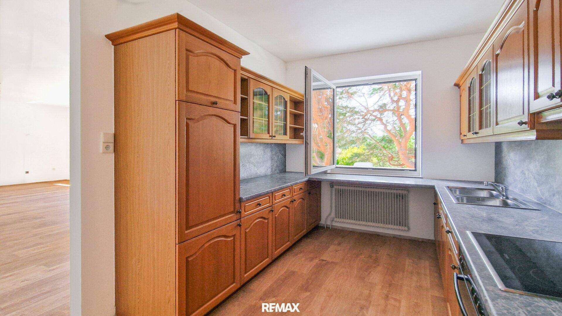Funktionale Küche mit Holzschränken, grauen Arbeitsplatten und einem Fenster mit Blick ins Grüne.