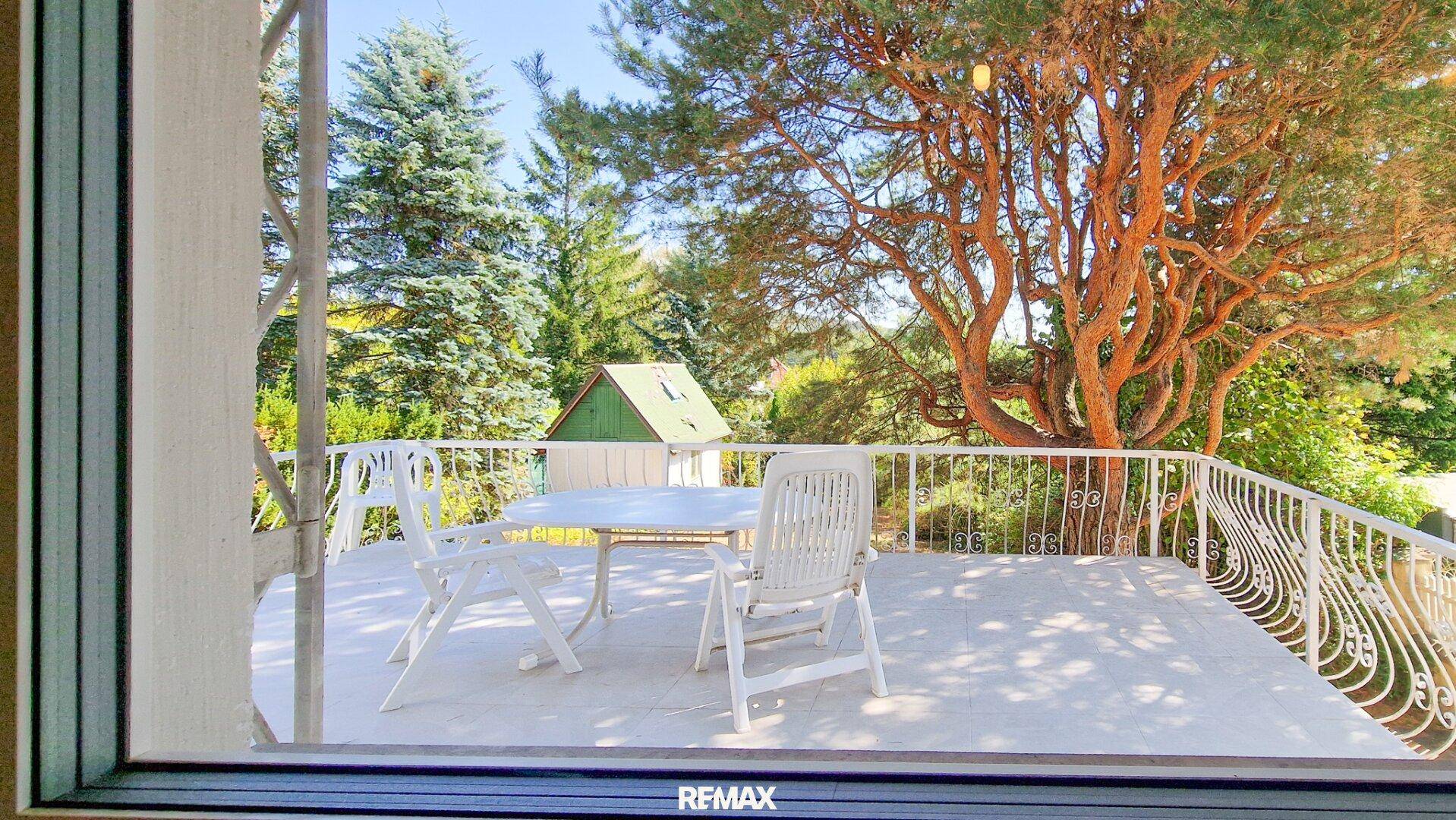 Blick aus dem Innenraum auf die sonnige Terrasse mit Gartenmöbeln und die umgebende grüne Landschaft.