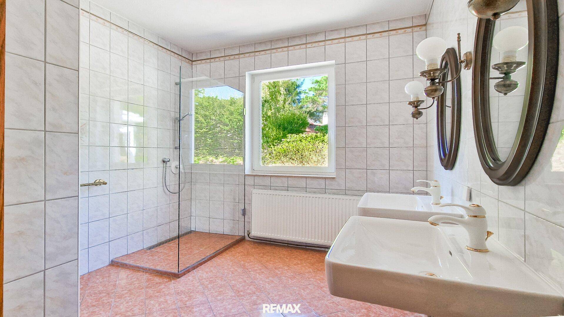 Helles Badezimmer mit zwei Waschbecken, begehbarer Dusche, Fenster und hellen Fliesen.