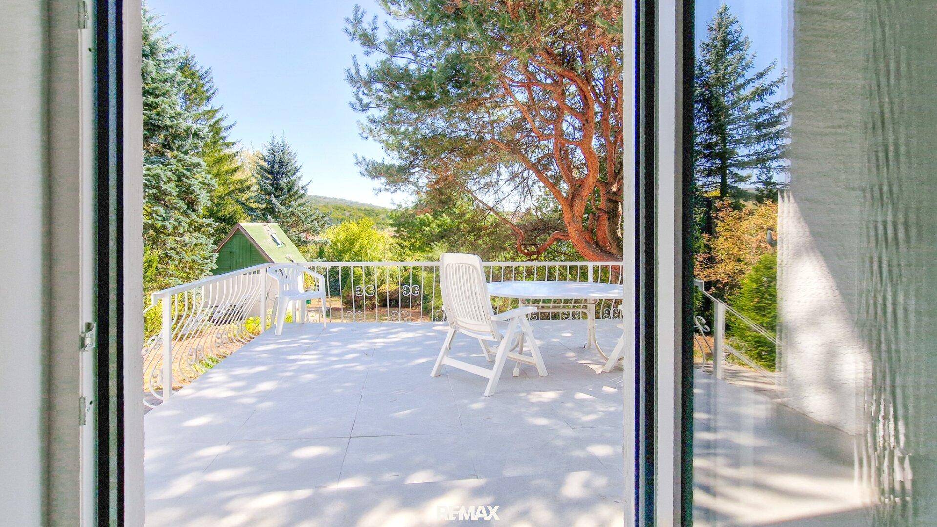 Blick aus dem Innenraum auf die sonnige Terrasse mit Gartenmöbeln und die umgebende grüne Landschaft.