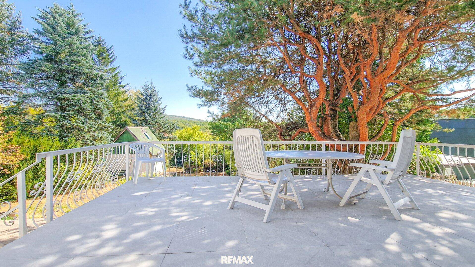 Sonnige Terrasse mit weißen Gartenmöbeln und schmiedeeisernem Geländer, umgeben von hohen Bäumen.
