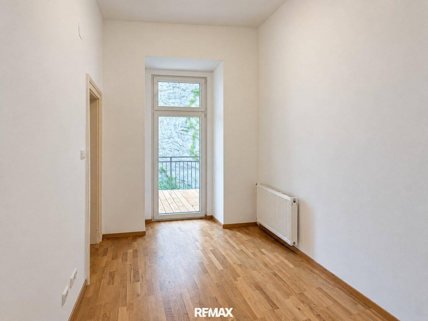 Leeres Zimmer mit Parkettboden und Zugang zu einem französischen Balkon mit Blick ins Grüne.