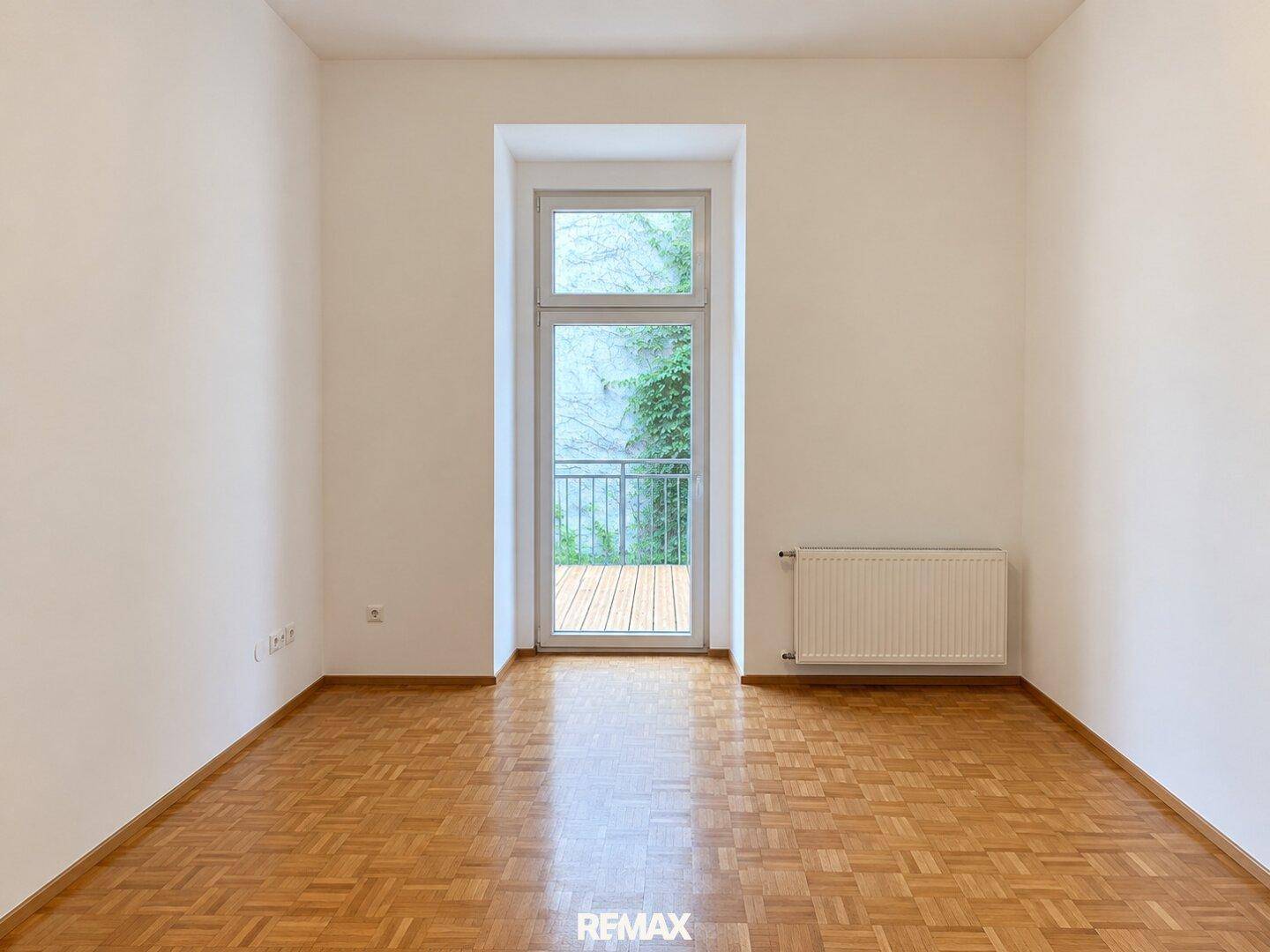 Helles Zimmer mit Holzboden und einem Fenster, das den Blick auf eine begrünte Wand freigibt.