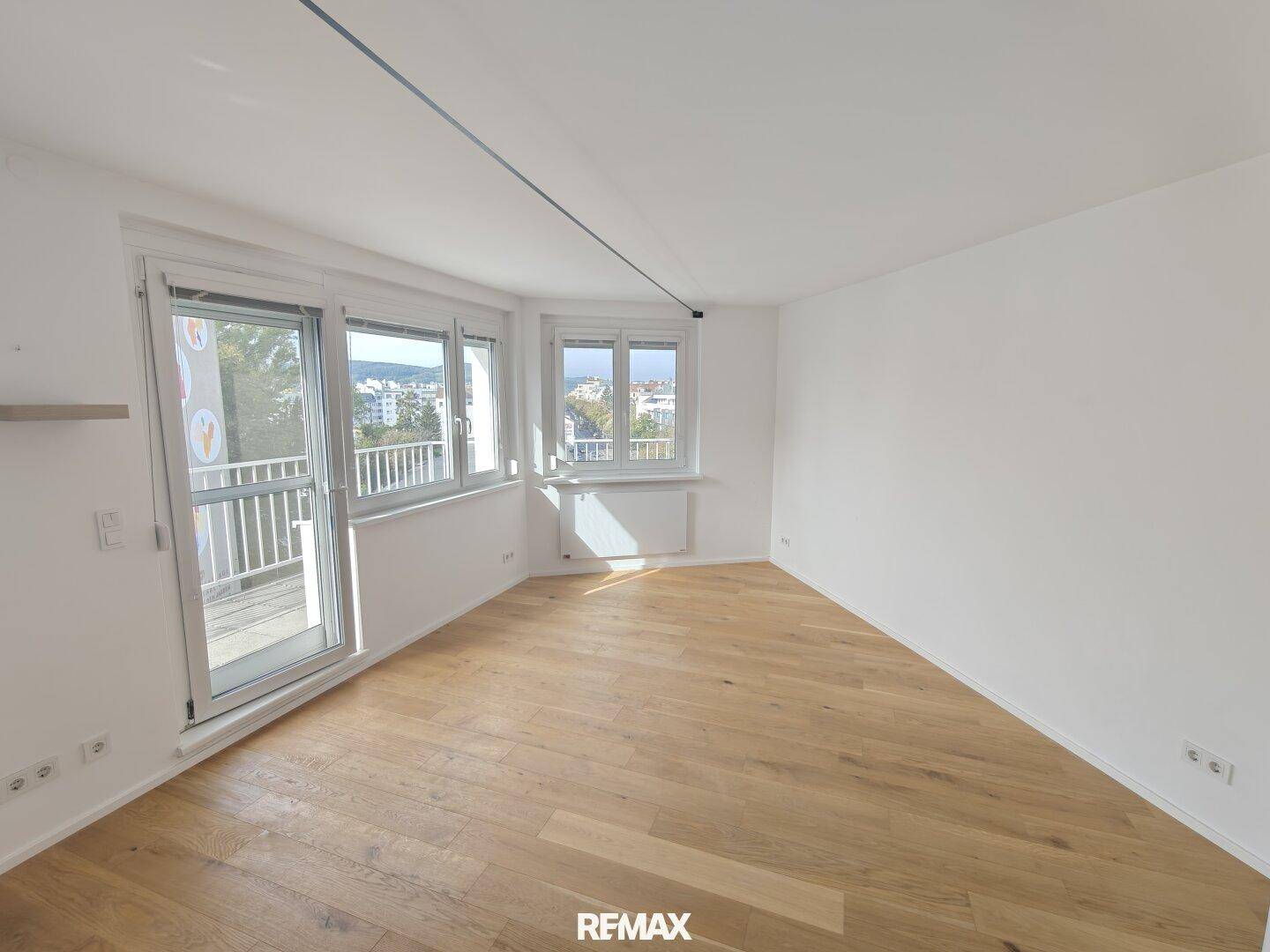 Heller, leerer Raum mit Holzboden, großen Fenstern und Zugang zu einem Außenbereich.