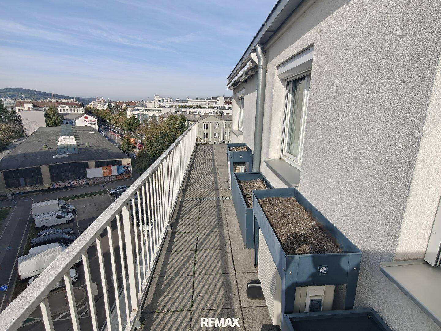Langer Balkon mit Stadtblick und Blumenkästen unter blauem Himmel.