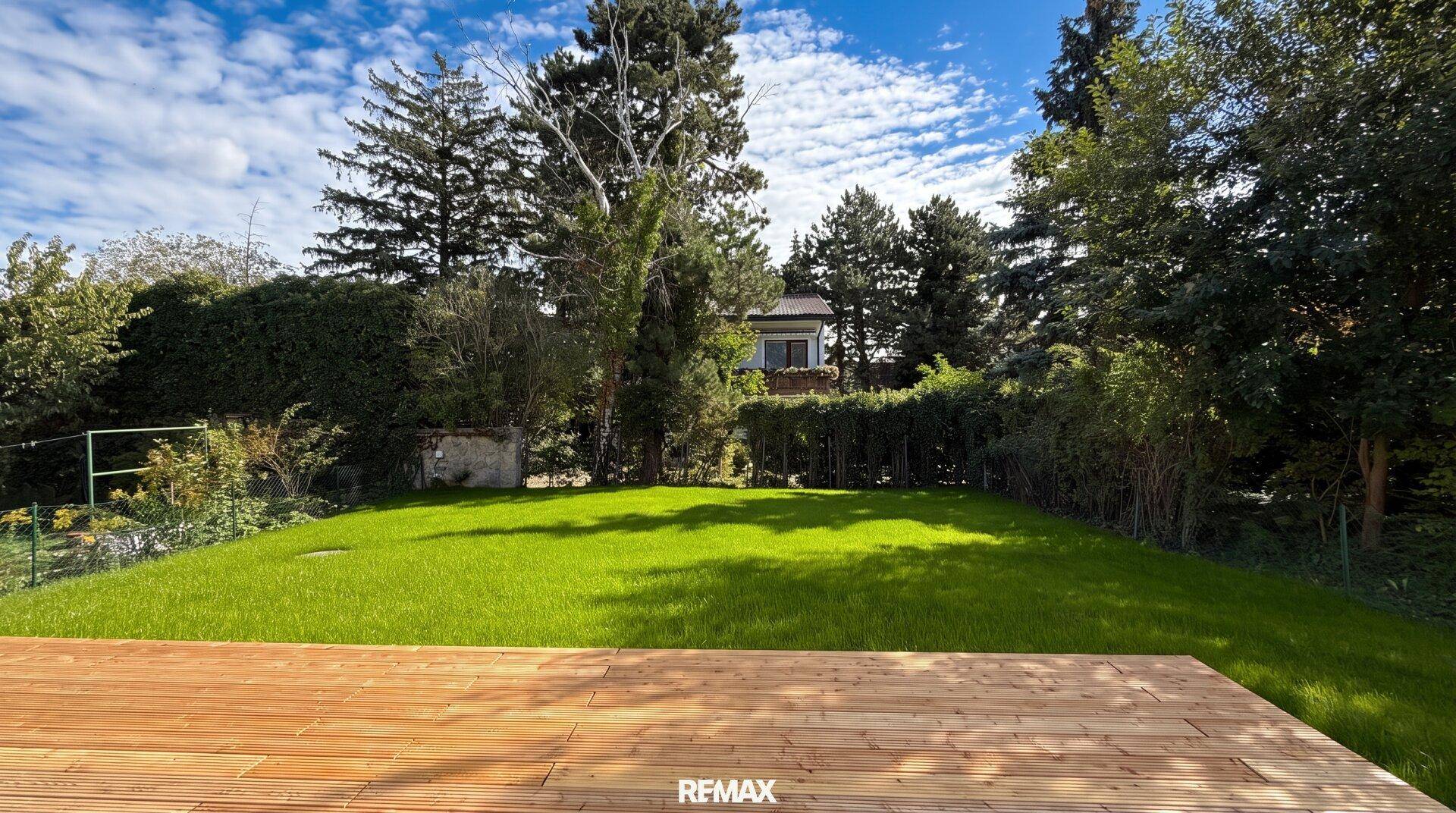 Großer, gepflegter Garten mit Rasenfläche und Holzterrasse unter blauem Himmel.