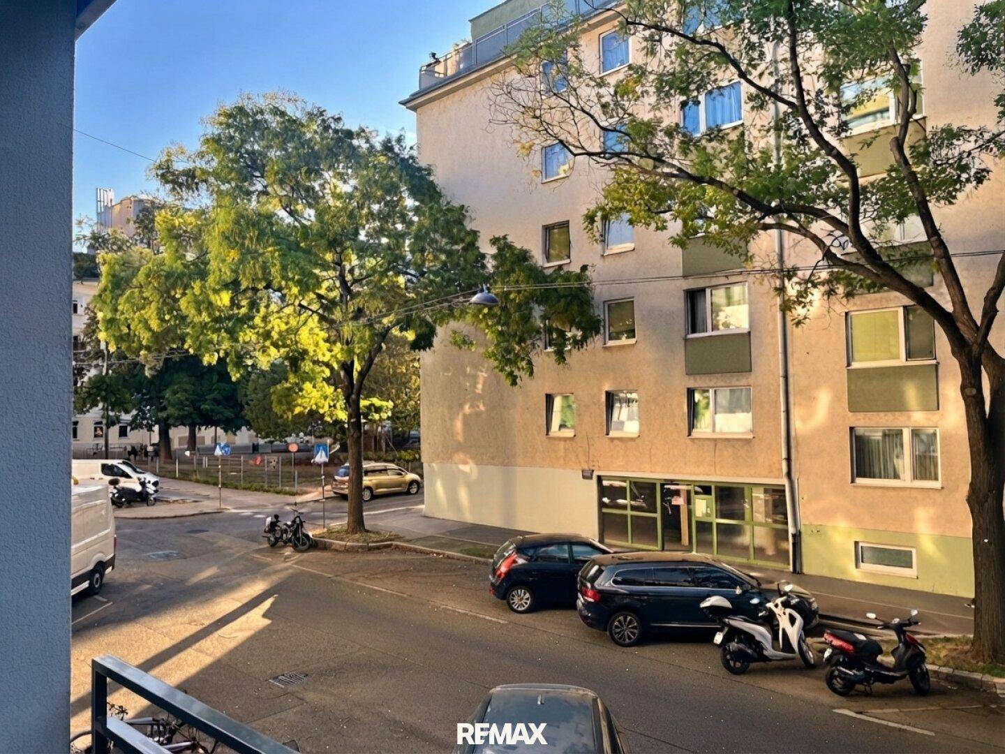 Blick von einem Balkon auf eine belebte Straße mit parkenden Autos und Bäumen.