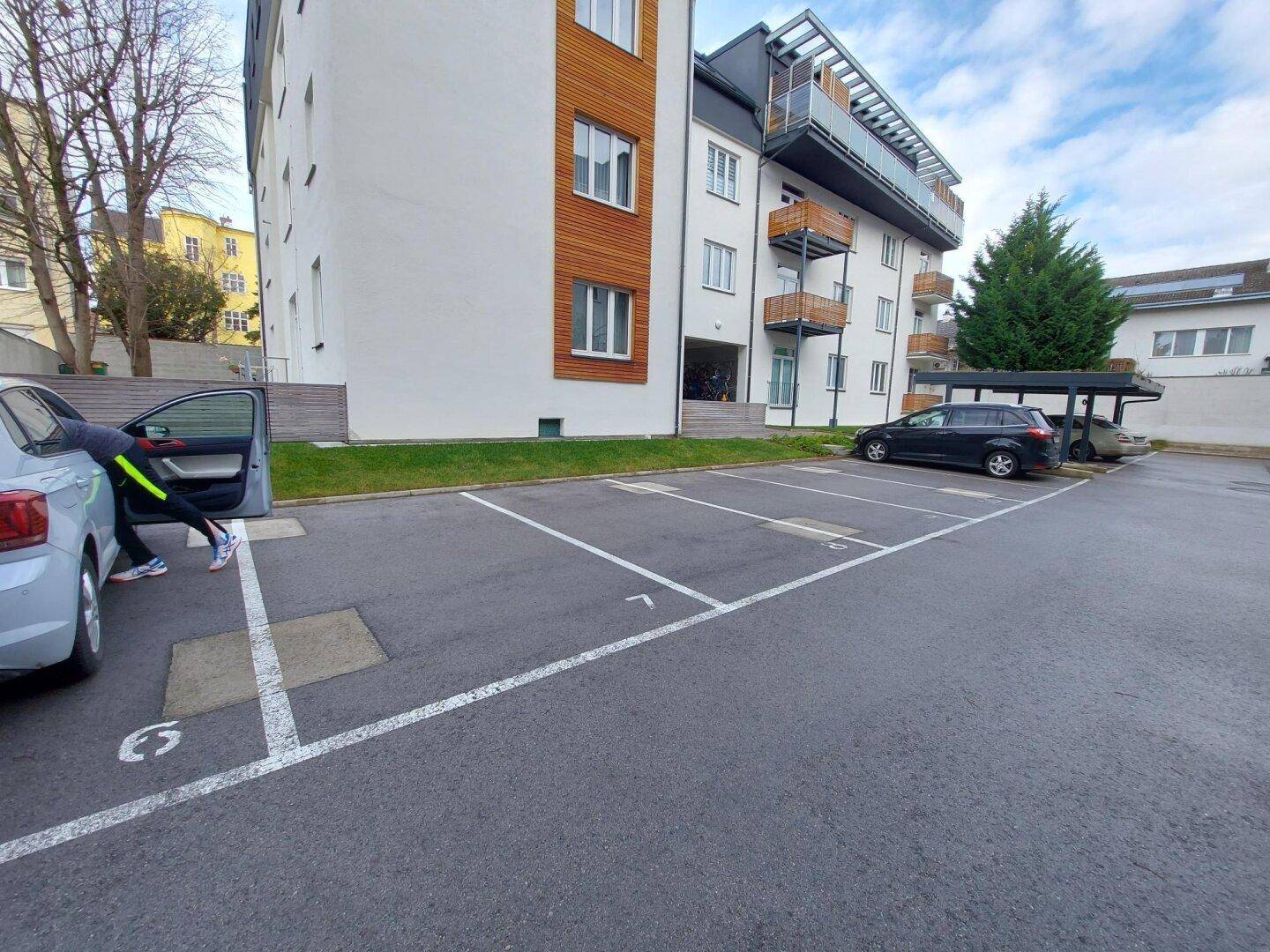 Wohngebäude mit moderner Fassade, Außenparkplätzen und einem Carport im Hintergrund.