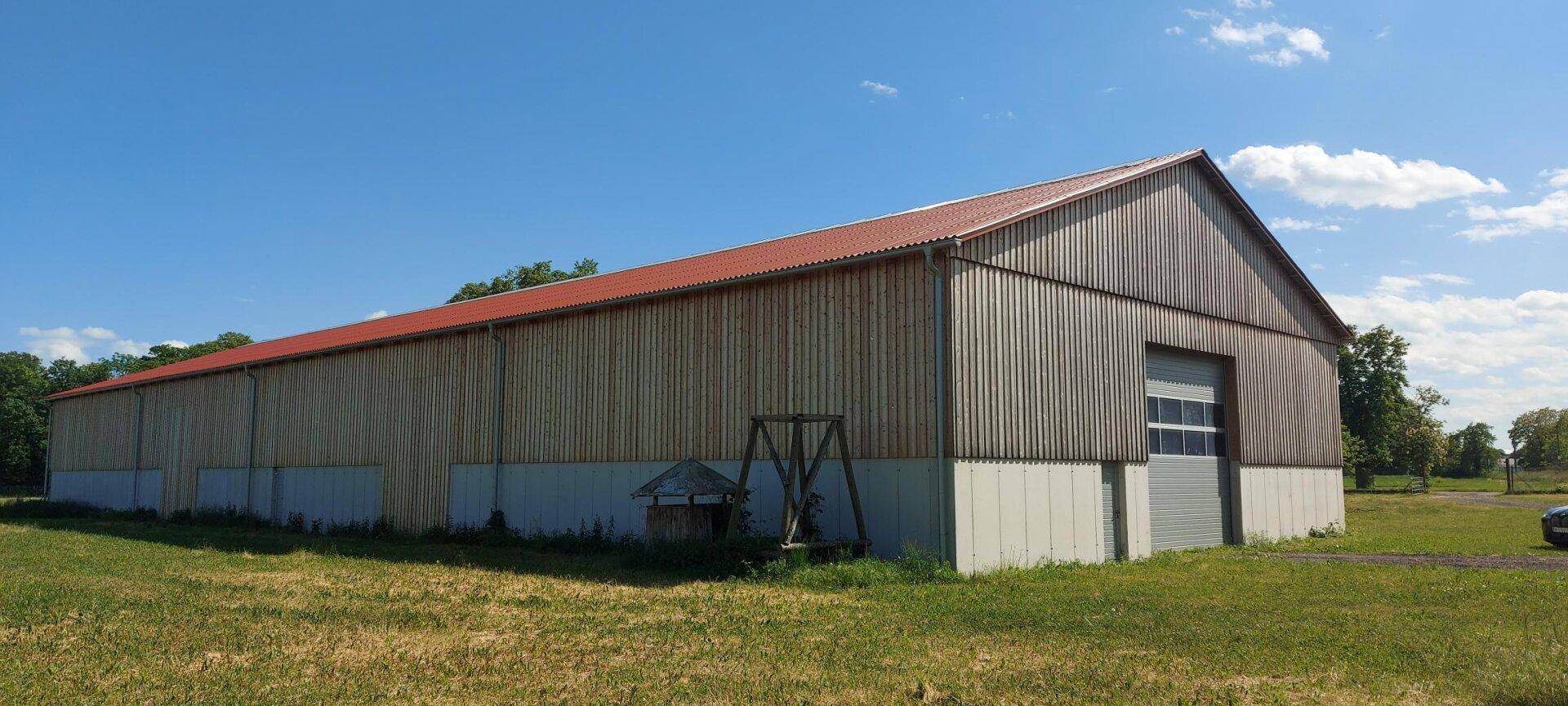 Ansicht der langen Lagerhalle mit Holzfassade, rotem Dach und weiten Grünflächen unter blauem Himmel.