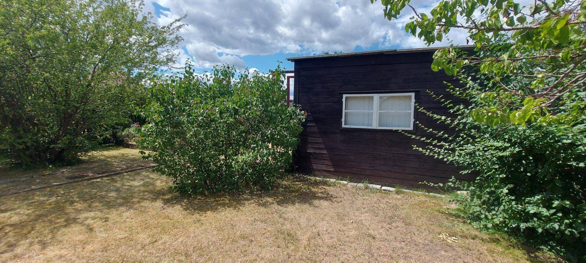 Kleines, dunkles Gartenhaus mit weißem Fenster, umgeben von üppigem Grün.