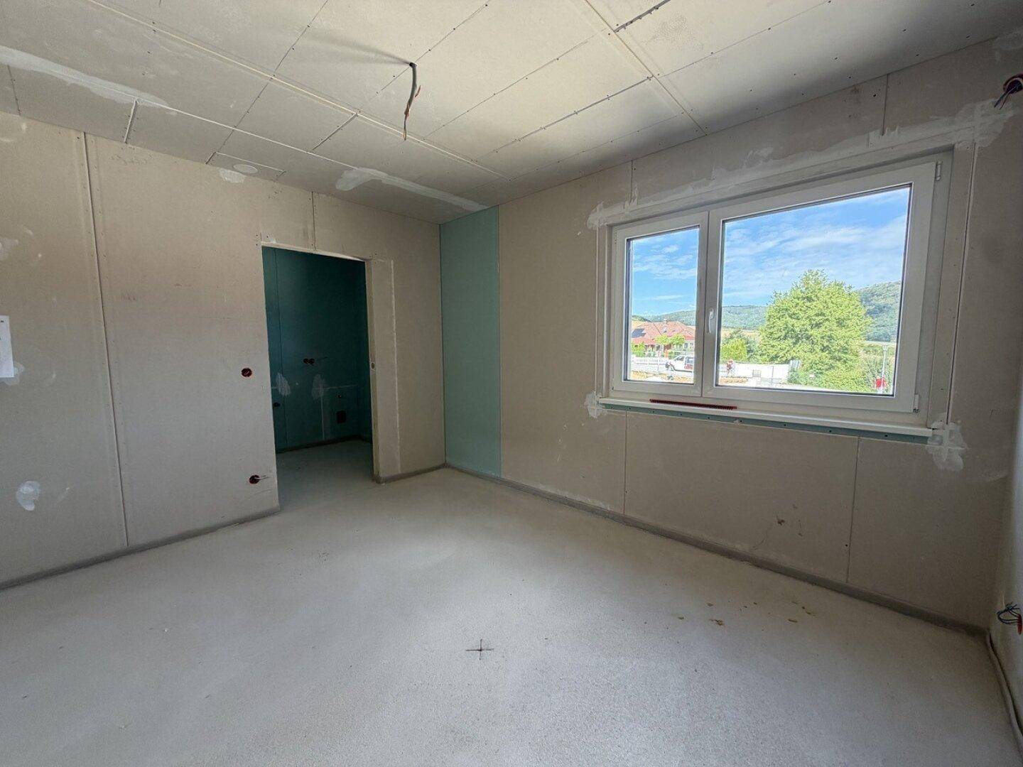 Helles Schlafzimmer im Rohbau mit großem Fenster und Ausblick auf die Umgebung.