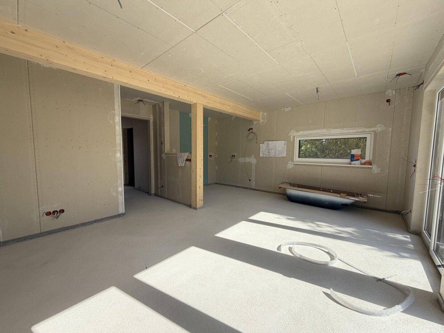 Geräumiger Wohnbereich im Rohbau mit sichtbaren Holzbalken und viel Tageslicht durch große Fenster.