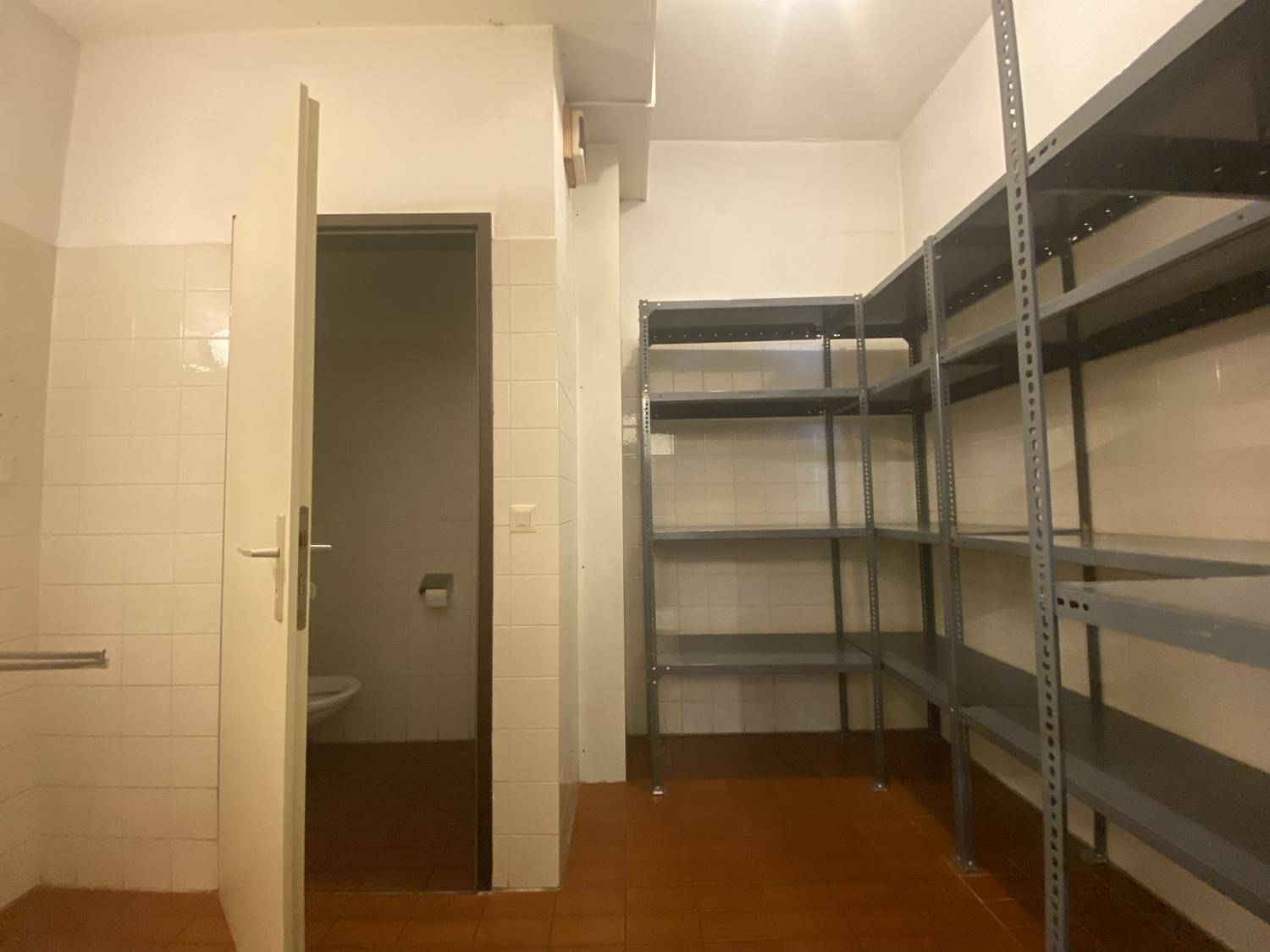 Kleiner Toilettenraum mit weißen Fliesen und angrenzendem Lagerbereich mit Metallregalen.