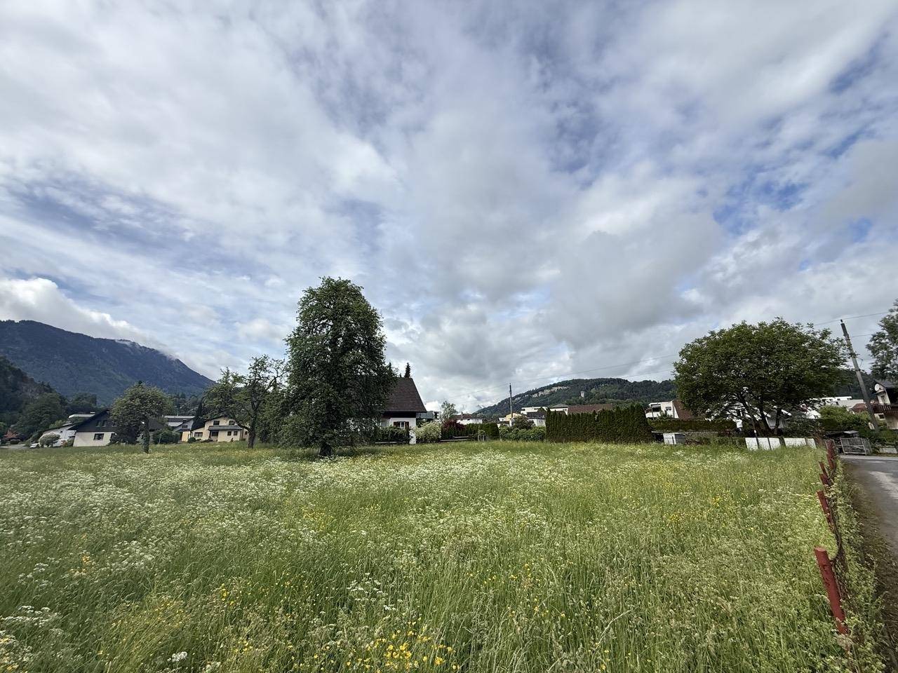 Weitläufige Wiesenlandschaft mit vereinzelten Häusern und einer beeindruckenden Bergkulisse am Horizont.