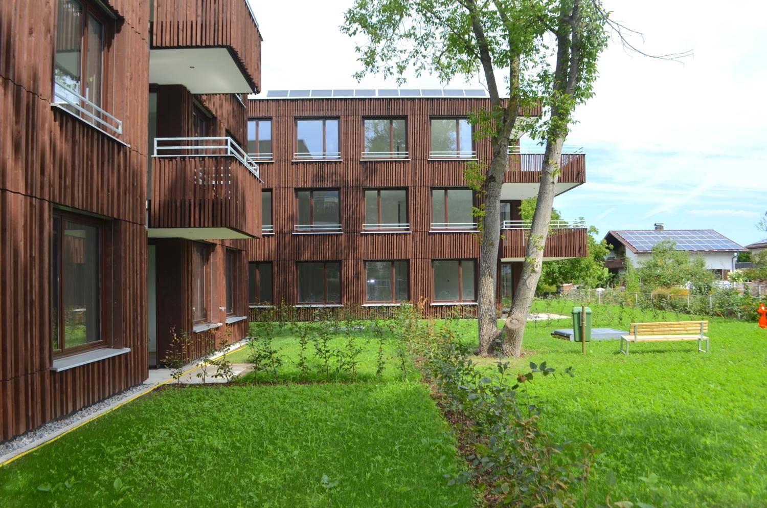 Grüne Gartenanlage vor dem modernen Gebäude mit Holzfassade, Balkonen und Sitzbank.