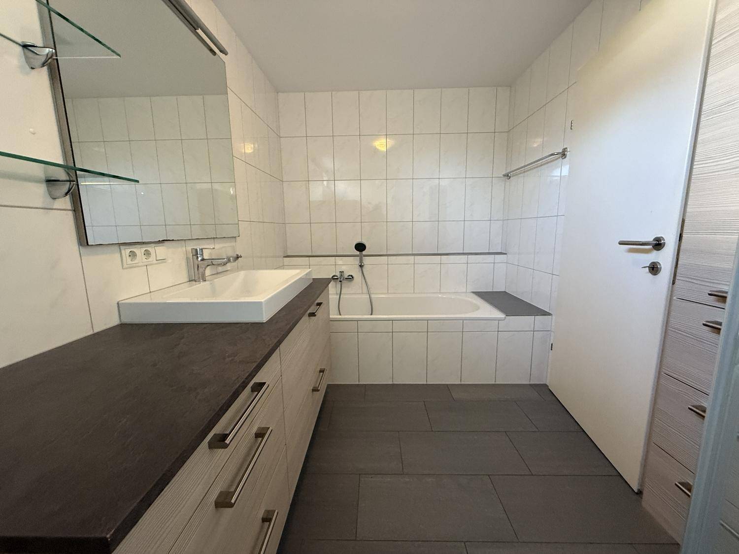 Helles Badezimmer mit Badewanne, Waschtisch und großem Spiegel, gefliest in Weiß und Grau.