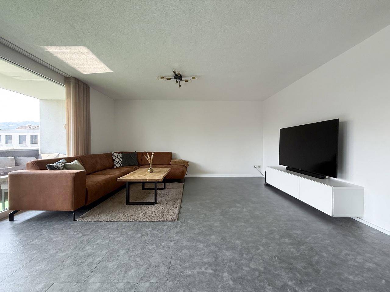 Gemütliches Wohnzimmer mit großem Sofa, Fernseher und Zugang zum Balkon.