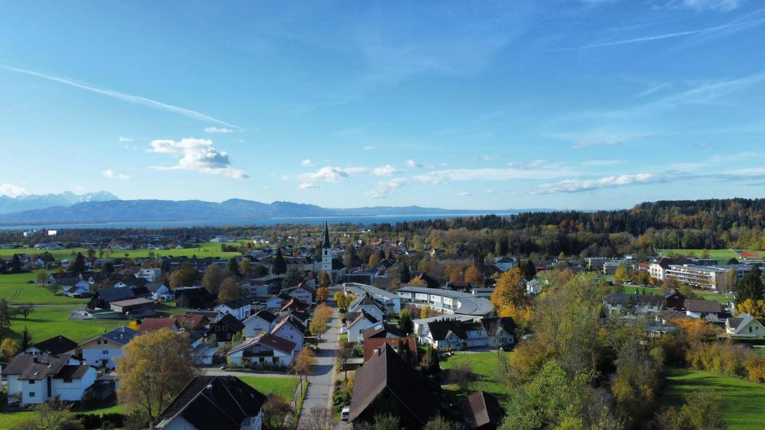 Herbstliche Luftaufnahme eines Dorfes mit See, Bergen und weiten grünen Flächen.