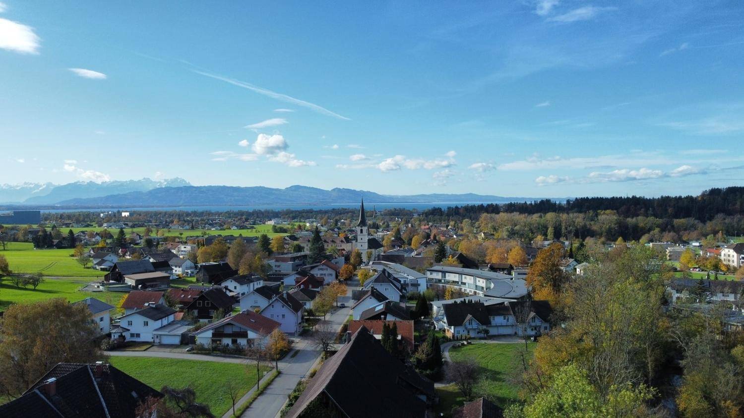 Weitläufige Luftaufnahme eines Dorfes mit Kirche, See und Alpenpanorama im Herbst.
