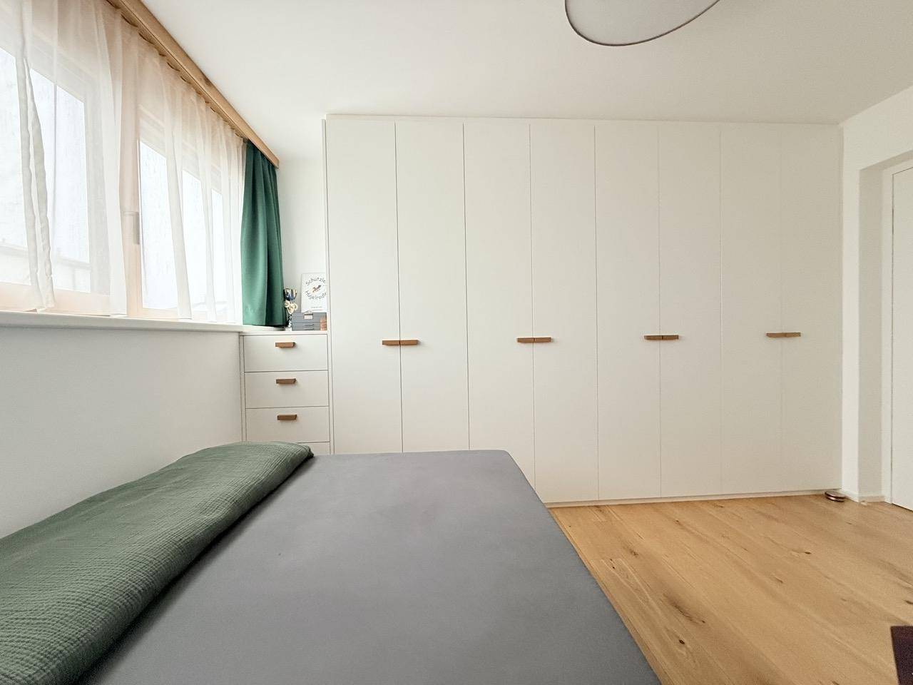 Helles Schlafzimmer mit Holzboden, Einbauschränken und Fenster mit Vorhängen.