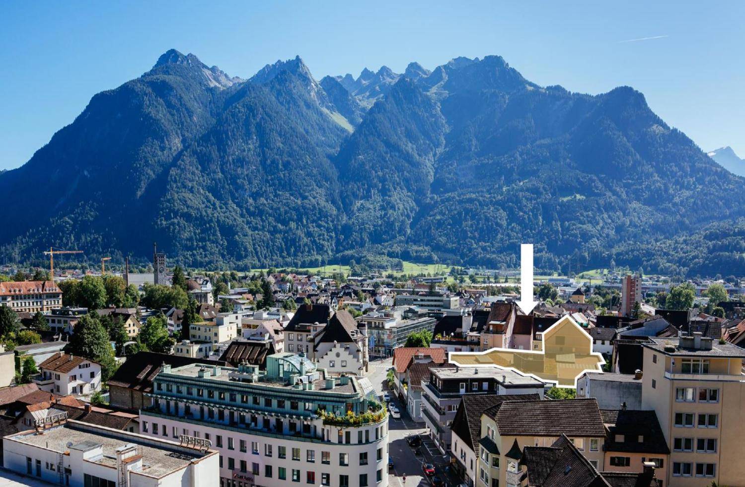 Luftaufnahme einer Stadt mit markiertem Gebäude vor einer beeindruckenden Bergkulisse unter blauem Himmel.