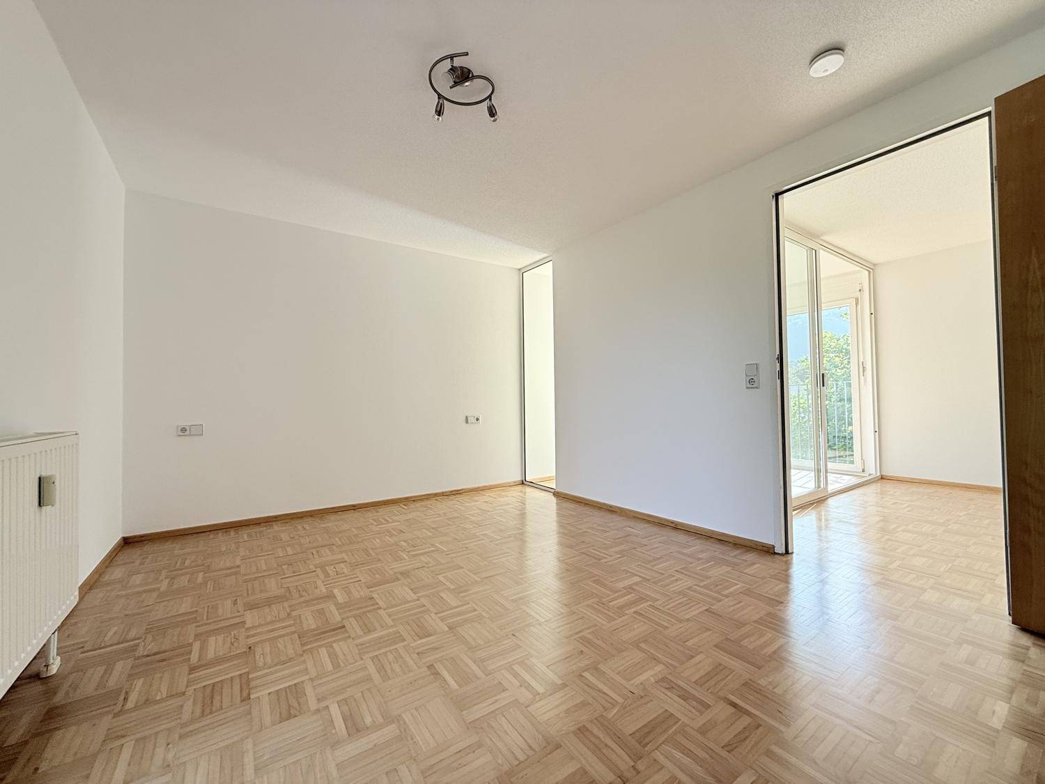 Heller Raum mit Parkettboden und Durchgang zu einem weiteren Zimmer mit großen Fenstern.