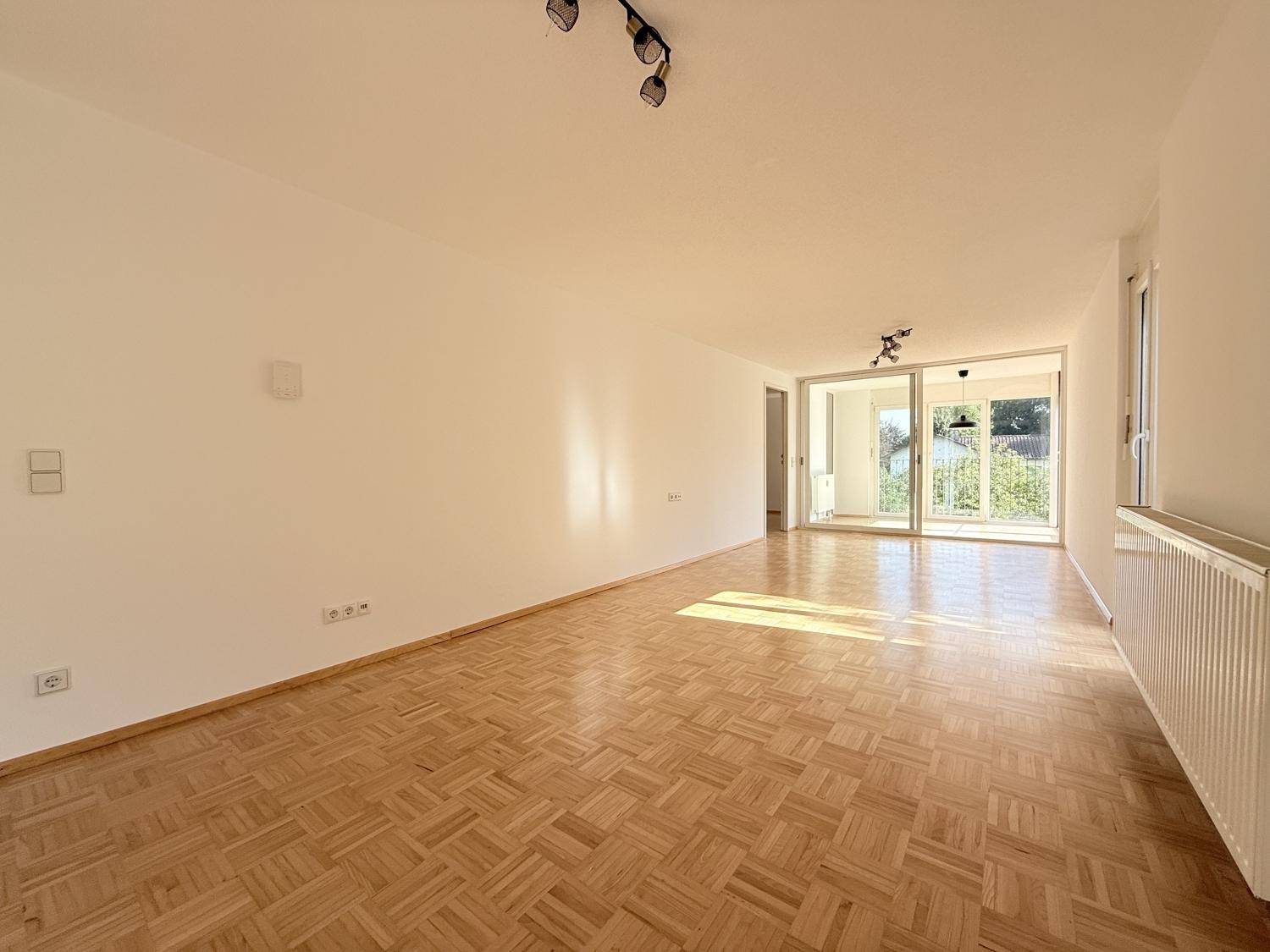 Helles, leeres Wohnzimmer mit Parkettboden und großen Schiebetüren zum Balkon.