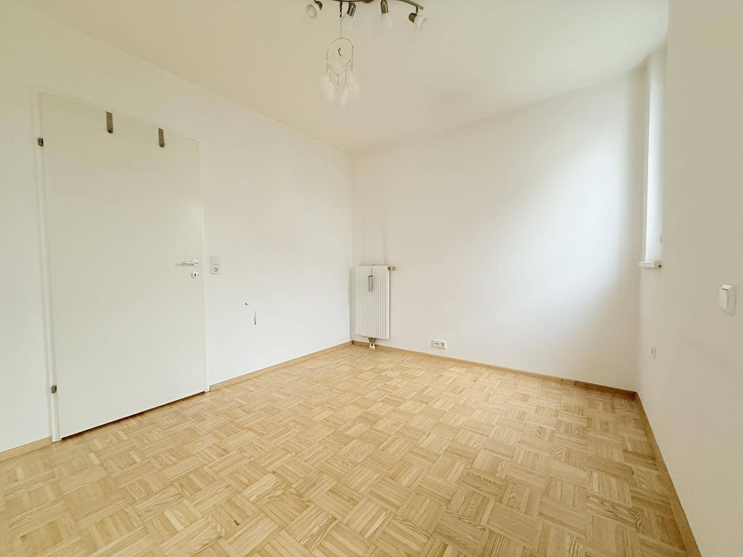 Leeres Zimmer mit hellem Parkettboden, weiß gestrichenen Wänden und einem Fenster.