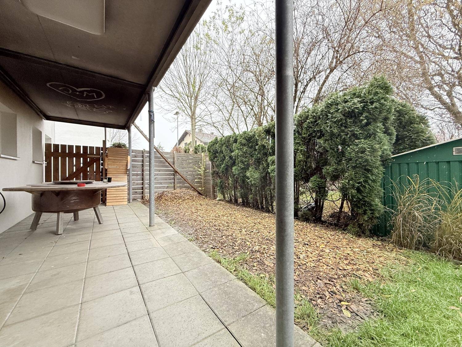 Überdachte Terrasse mit Sitzgelegenheit und Blick auf den Garten mit Hecken und Geräteschuppen.