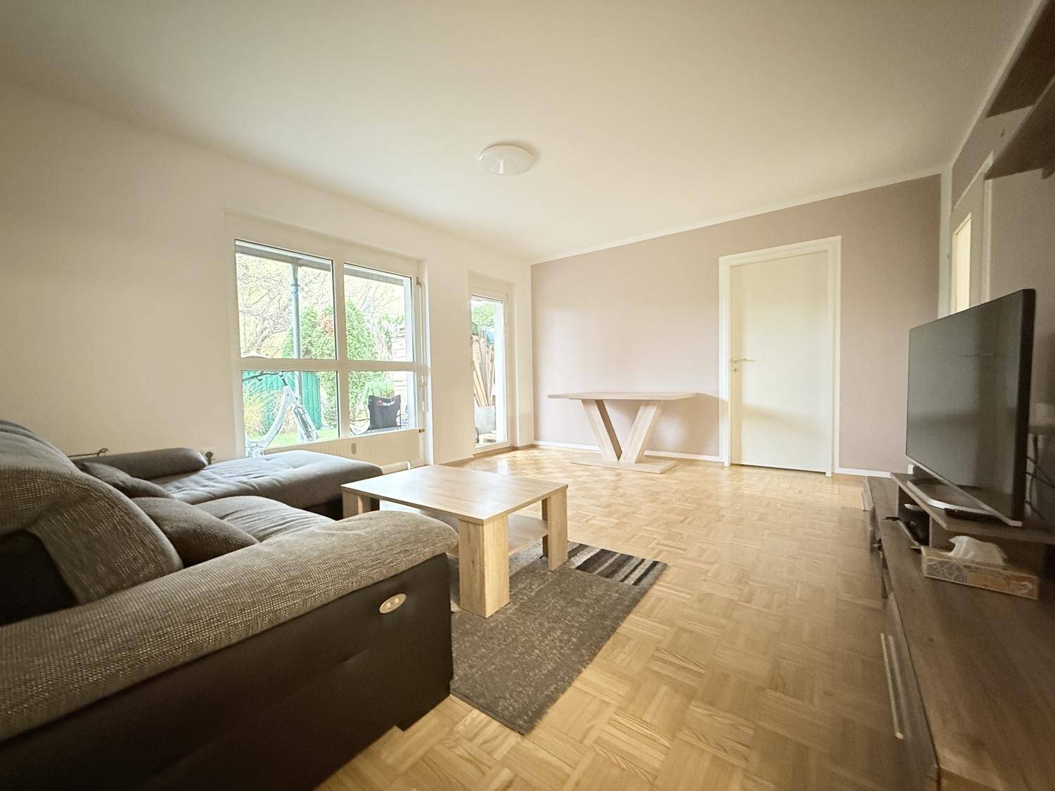 Helles Wohnzimmer mit Parkettboden, großem Fenster und Zugang zu einem Außenbereich.