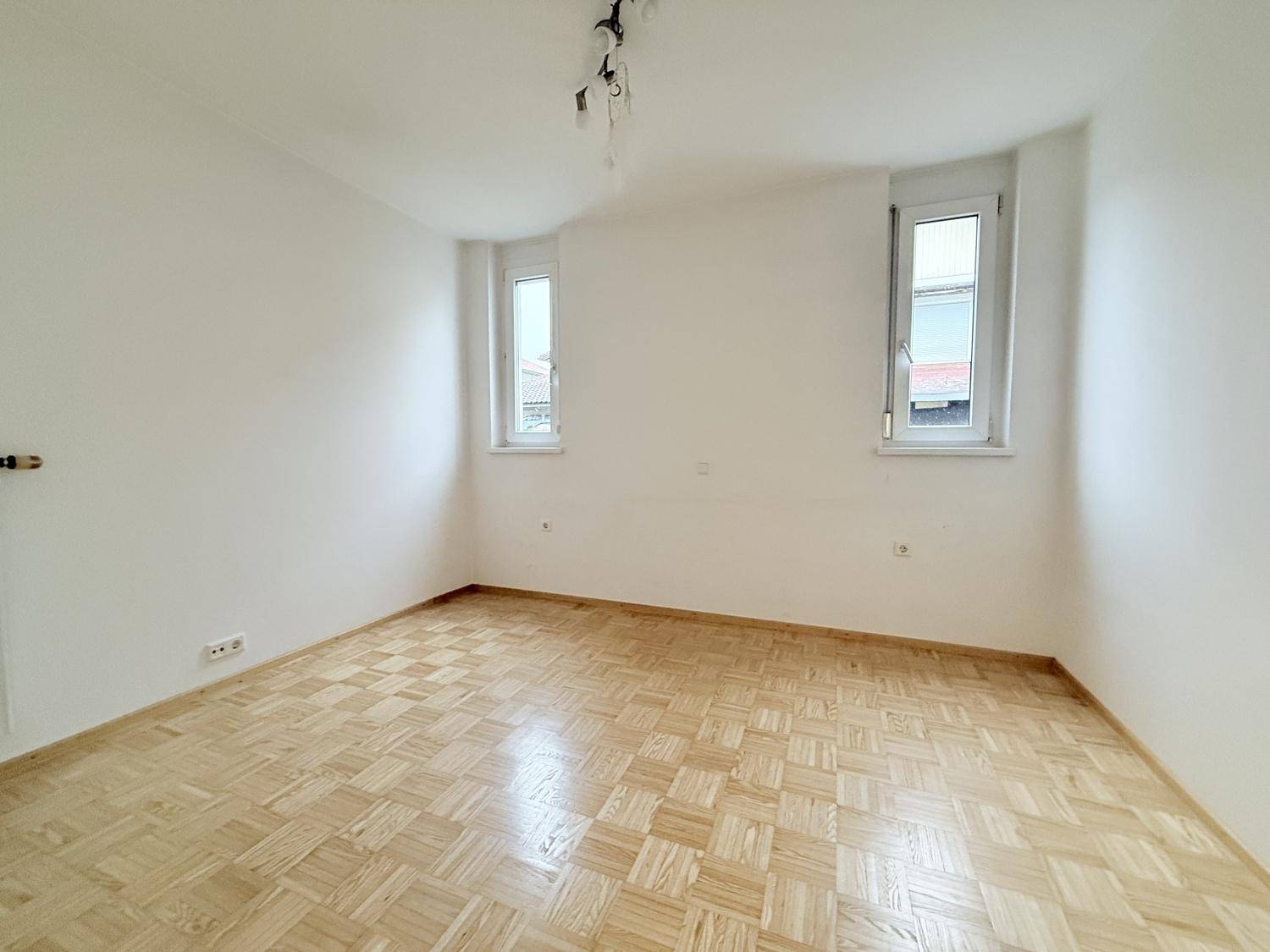 Helles, leeres Zimmer mit Parkettboden und zwei Fenstern, ideal als Schlaf- oder Arbeitszimmer.