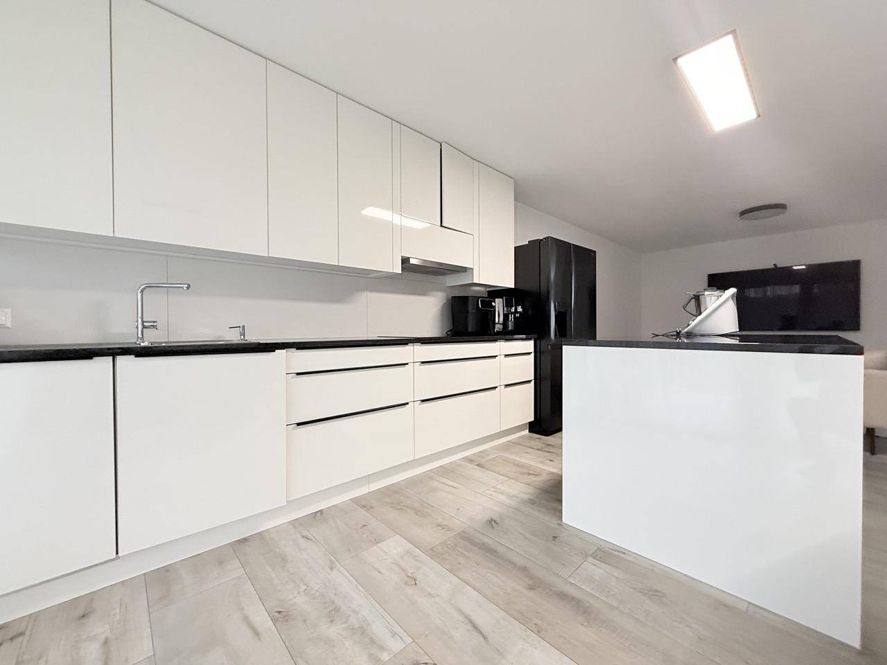 Moderne weiße Küche mit Kochinsel, schwarzen Arbeitsplatten und integriertem Kühlschrank.
