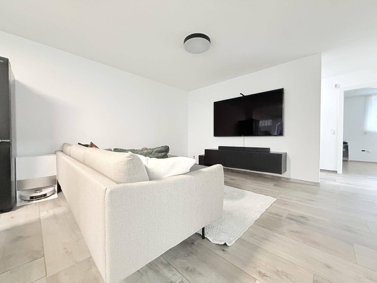 Heller Wohnbereich mit beigem Sofa, Couchtisch und großem Wandfernseher.