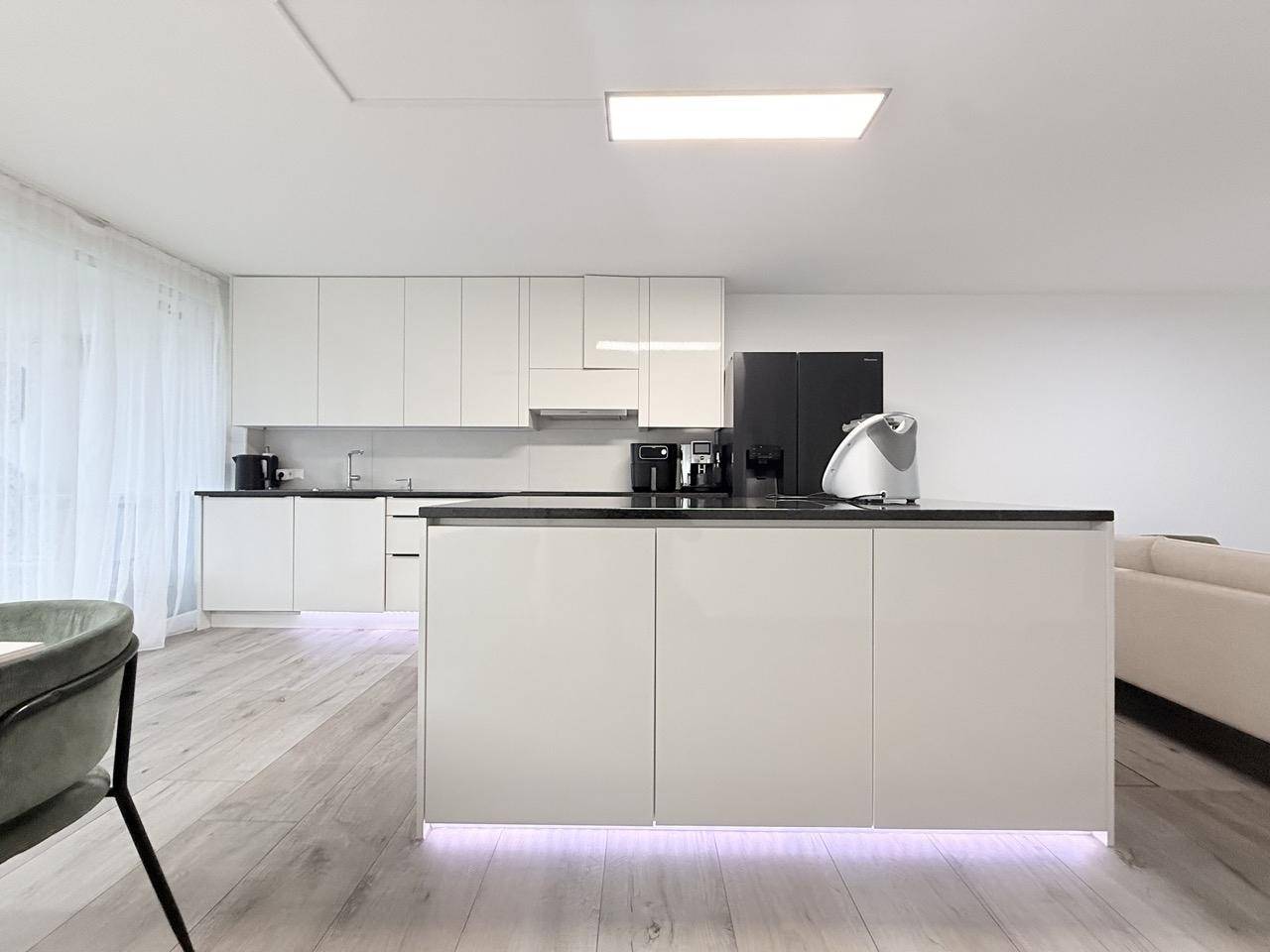 Moderne weiße Küche mit Kochinsel, schwarzen Arbeitsplatten und integrierten Geräten.