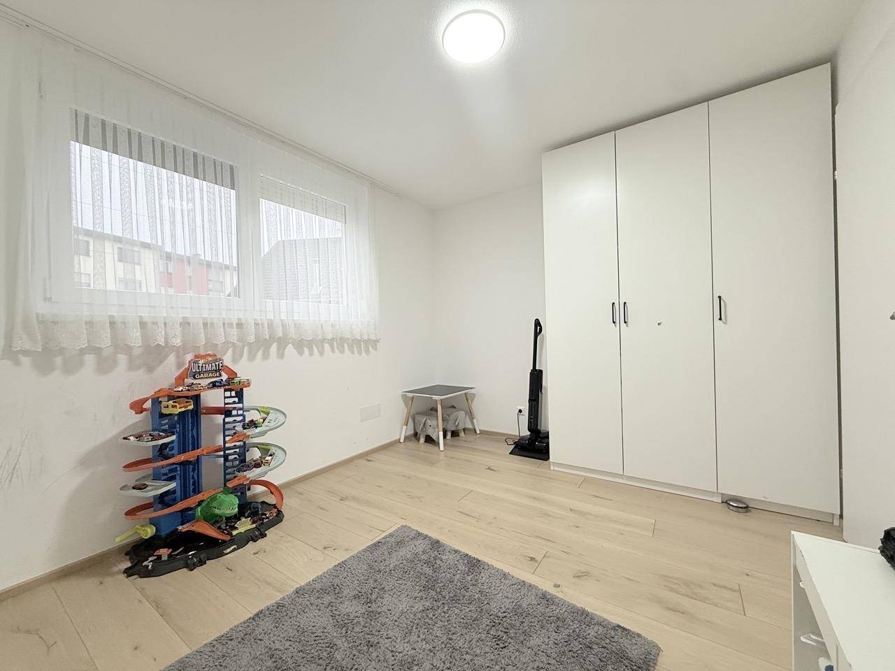 Kinderzimmer mit hellem Holzboden, Fenster und weißem Kleiderschrank.