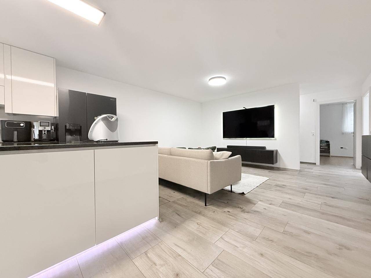 Heller Wohnbereich mit großem Fernseher, Sofa und Blick auf die offene Küche.