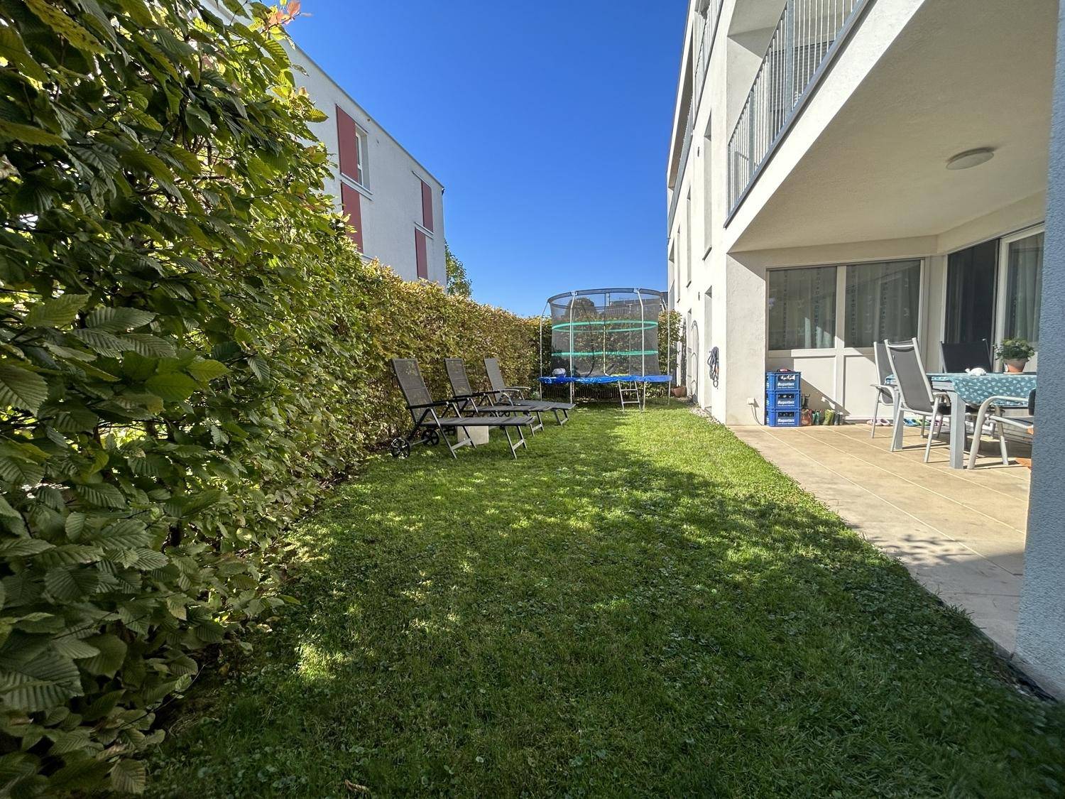 Gepflegter Gartenbereich mit Rasenfläche, Hecke, Sonnenliegen und Trampolin neben der Terrasse.