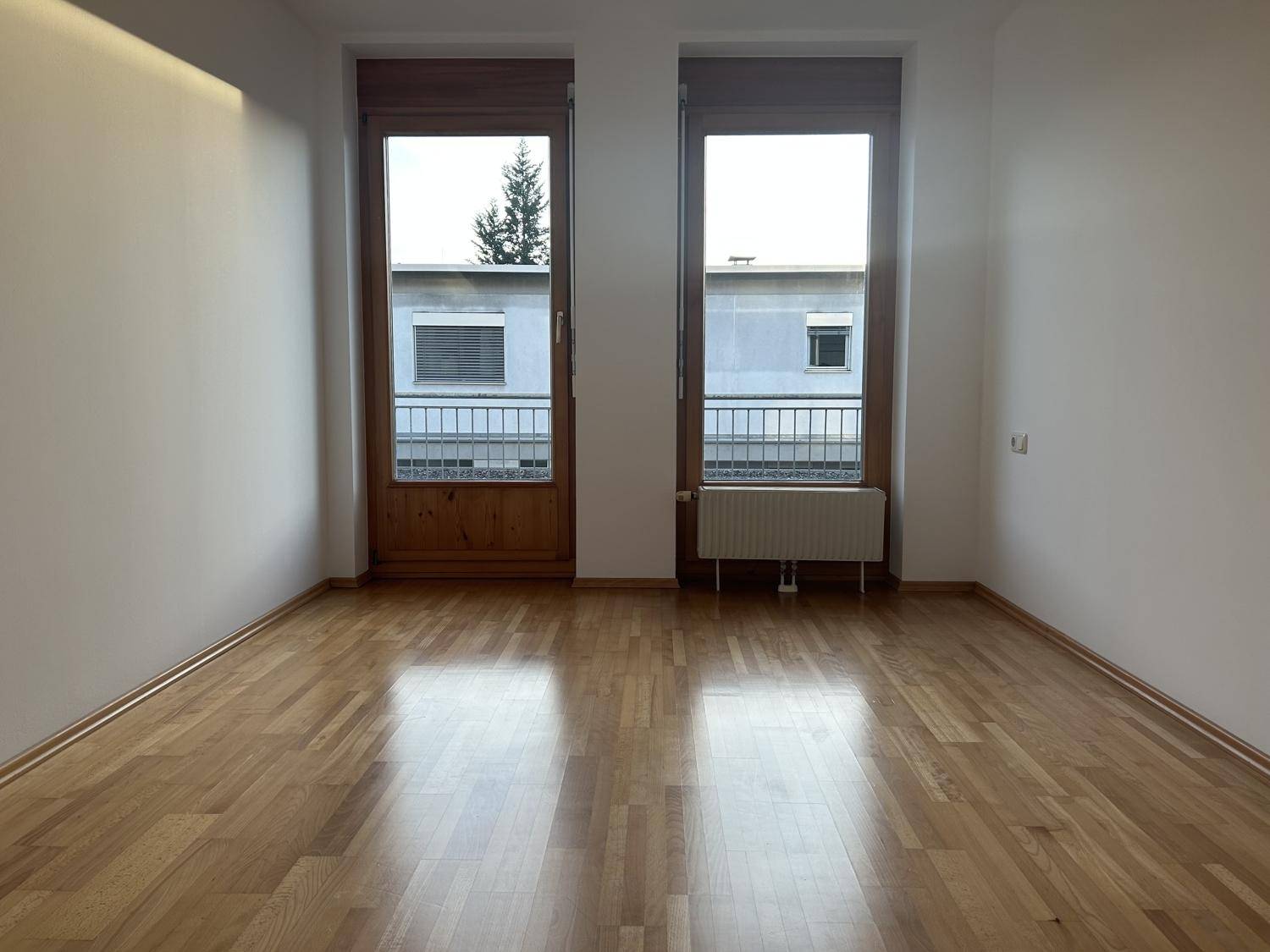 Helles Zimmer mit Holzparkettboden und zwei Fenstertüren, die zu einem französischen Balkon führen.